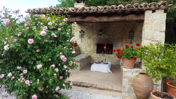 Un espace extérieur aménagé avec des sièges en pierre, doté d’un foyer, et entouré de plantes en pot et de rosiers en fleurs.