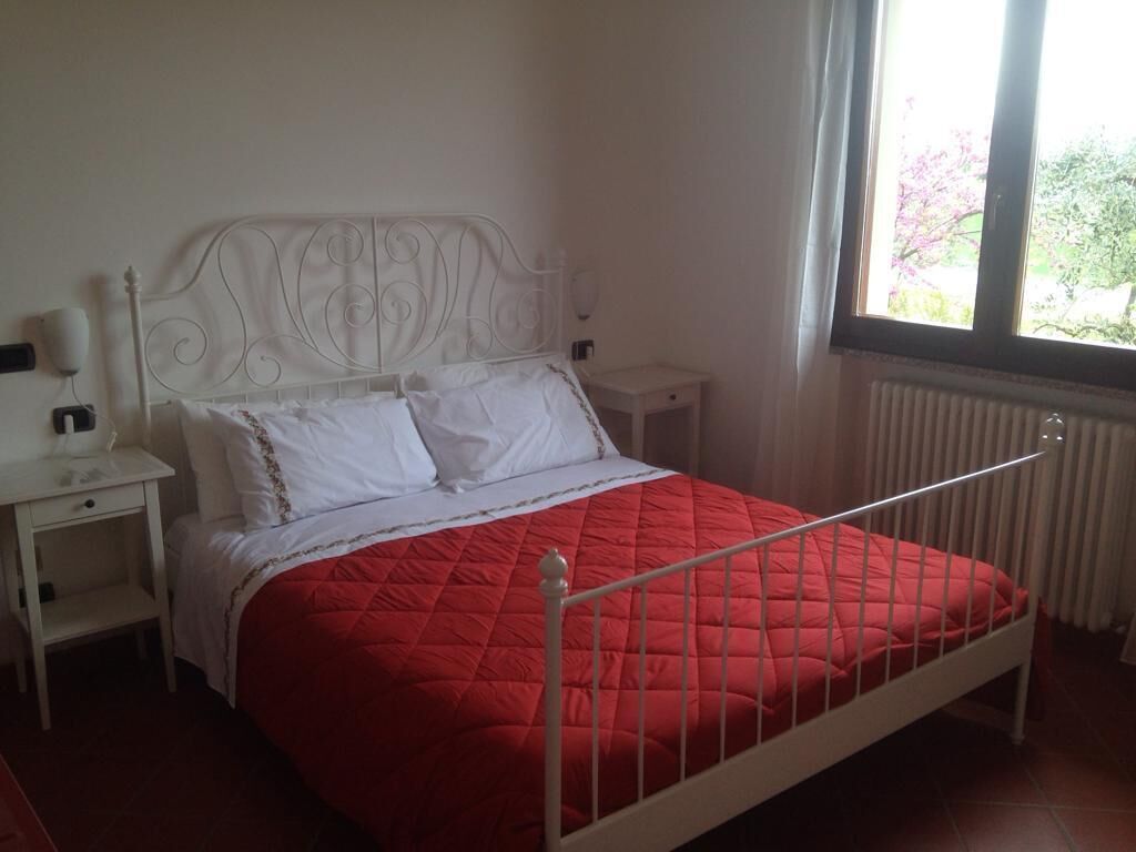 Un letto con struttura in metallo bianco e copripiumino trapuntato rosso, un comodino bianco con una lampada e una finestra con tende.