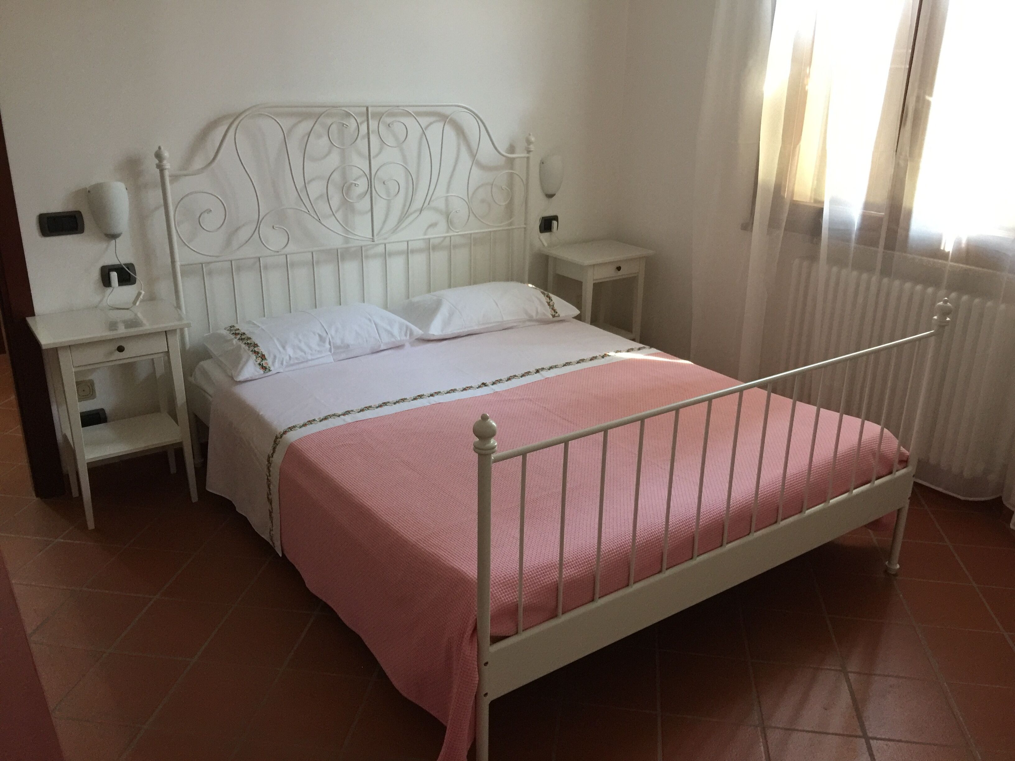 A bed with a metal frame, white bedding, and a pink bedspread. There are two bedside tables with lamps and a wall-mounted light.