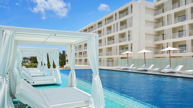 A pool area with white canopied loungers and umbrellas, a clear blue pool, and a multi-story building with balconies.