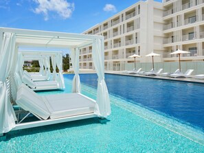 Un área de piscina con tumbonas blancas cubiertas por toldos y sombrillas, una piscina azul cristalina y un edificio de varios pisos con balcones.
