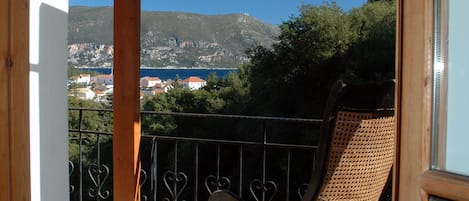 A wooden balcony with a wicker rocking chair, stone flooring, and a view of a coastal town and mountains.