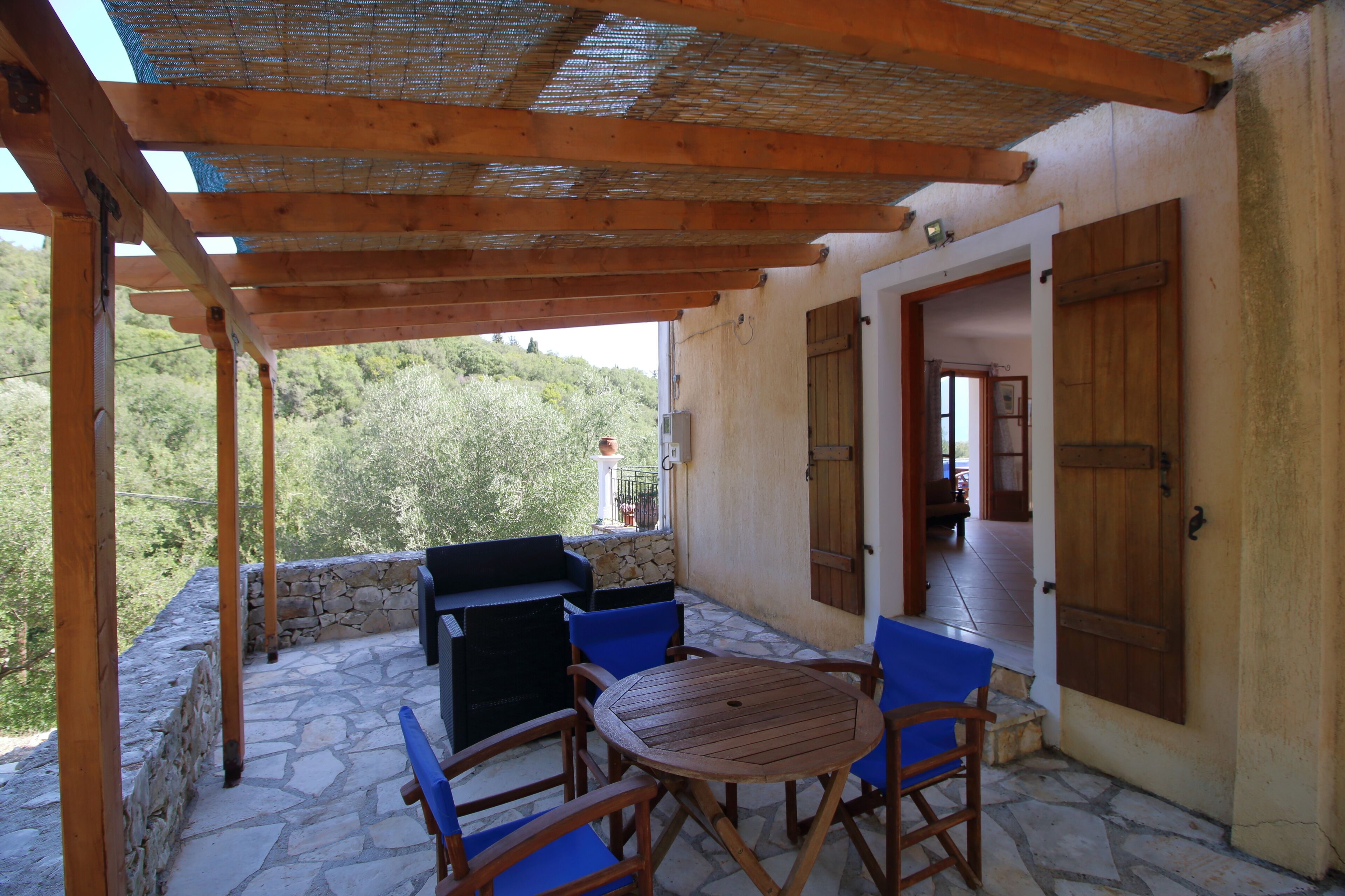 A covered patio with stone flooring, wooden furniture, and a view of greenery.