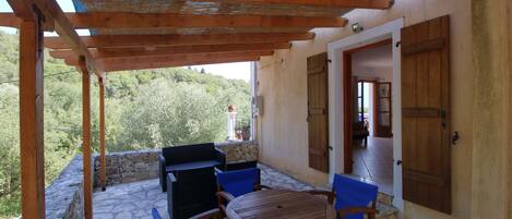 A covered patio with stone flooring, wooden furniture, and a view of greenery.