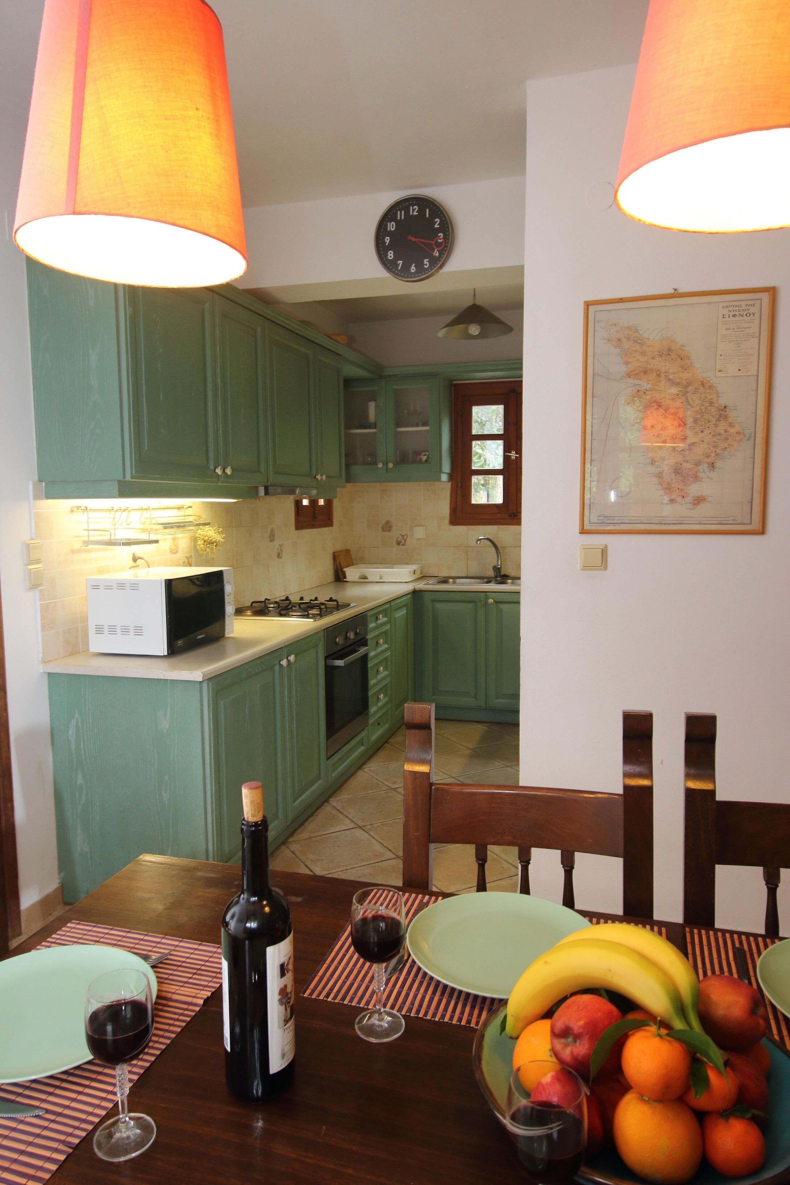A kitchen with green cabinets, a microwave, a stove, and a sink. There is a clock on the wall and a map hanging above the sink.