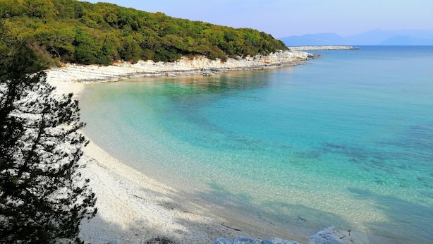 Eine Küstenlandschaft mit klarem türkisblauem Meer, weißem Sandstrand und einem bewaldeten Hügel.