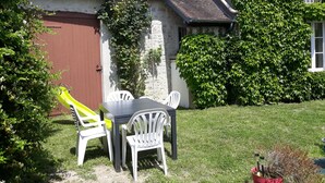 A rustic stone building with a brown door and a window. There is a white plastic table and chairs, a yellow slide, and a red flower pot with a plant. - Gîte Suzanne, in Essonne Verte, near Milly la Forêt (Brouy)