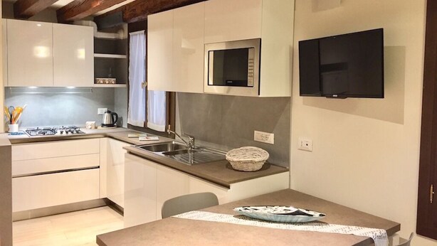 Una cocina compacta con armarios blancos, microondas, estufa y pileta. Hay un televisor de pantalla plana montado en la pared y una mesa con sillas.