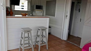 Una cocina compacta con gabinetes blancos, una pequeña isla con taburetes, un microondas y un ventanal con persianas.