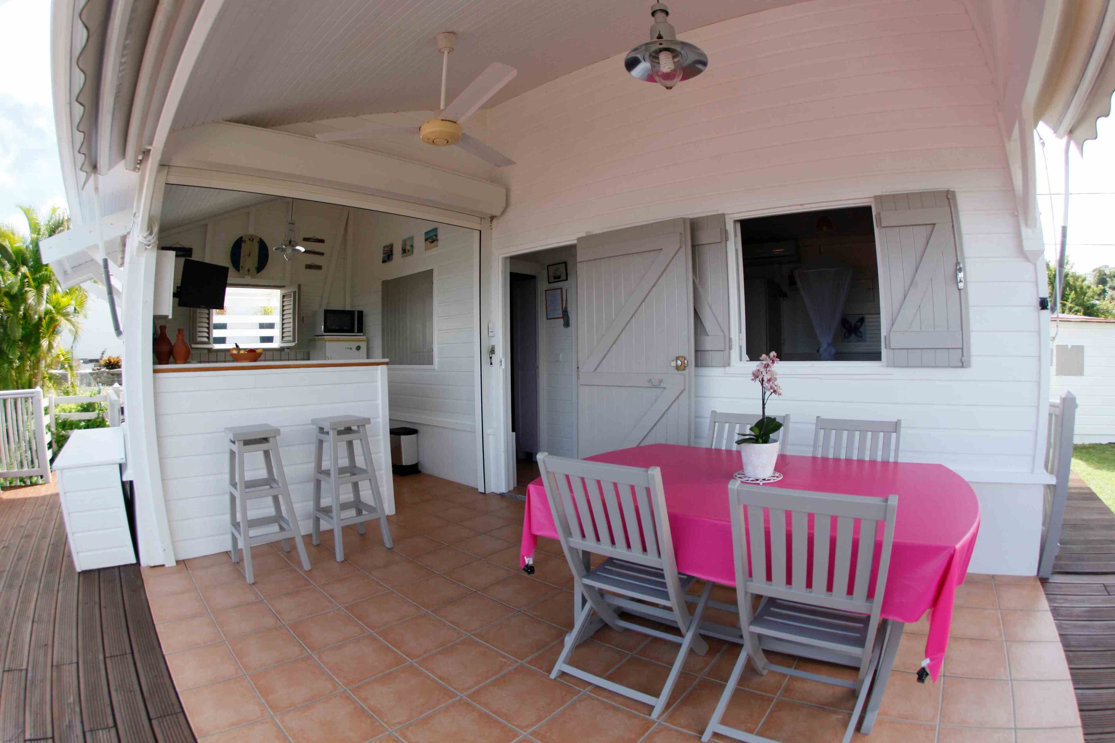 Un patio cubierto con una mesa, sillas, un ventilador de techo y una cocina visible a través de una puerta abierta.