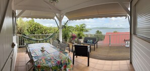 A covered outdoor patio with a table and chairs, a hammock, and a view of the sea and greenery.