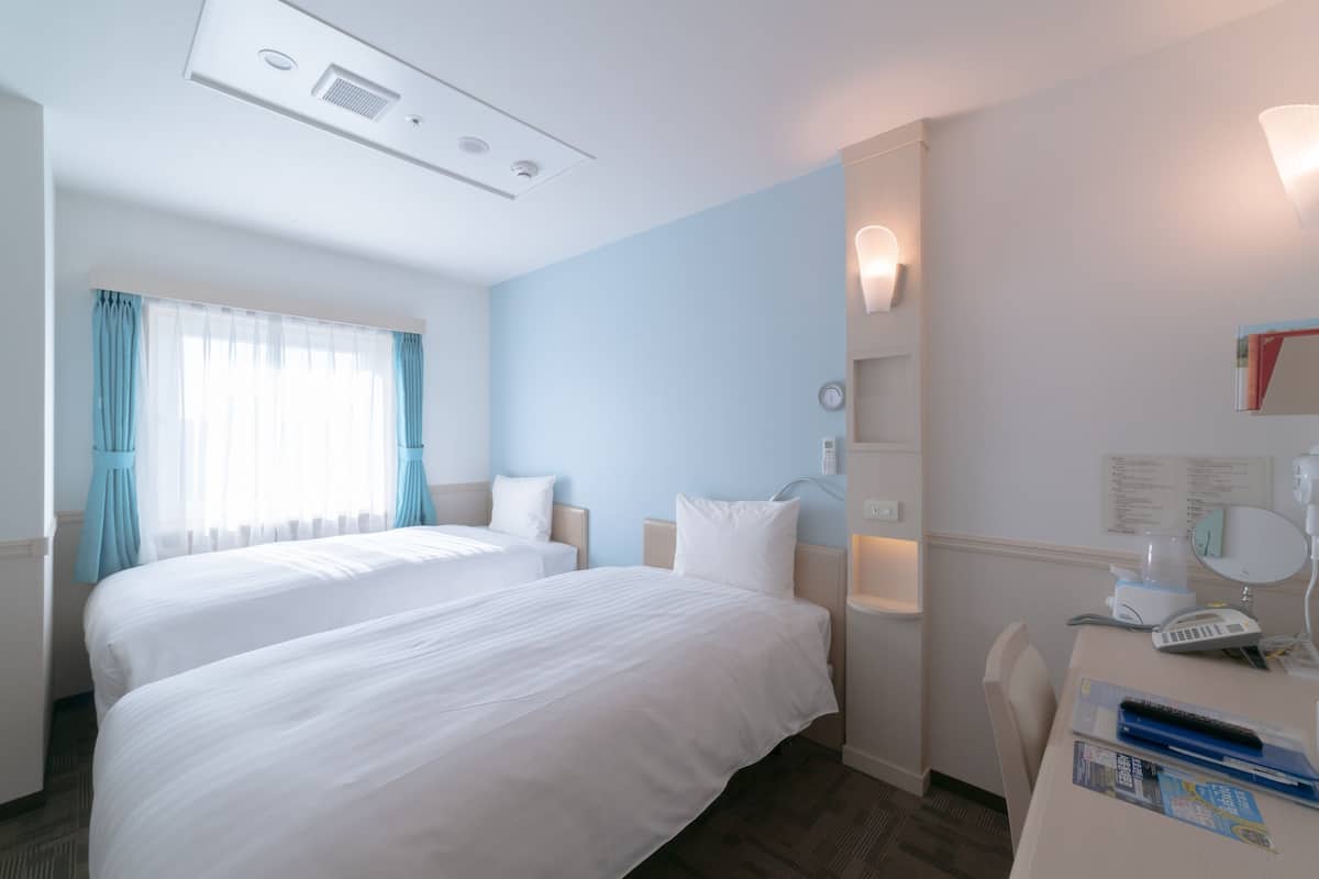 Une chambre d’hôtel avec deux lits, un bureau doté d’un téléphone et d’un miroir, une fenêtre avec des rideaux et un luminaire fixé au mur.