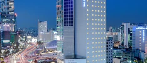 A modern hotel with a prominent 'HOTEL' sign, located at a busy intersection with multiple lanes of traffic and surrounding high-rise buildings.
