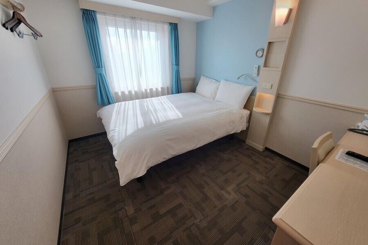 Une chambre d’hôtel avec un lit, un bureau, une chaise, une fenêtre avec des rideaux et une horloge murale.