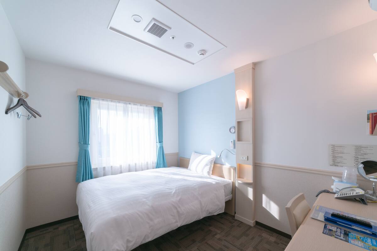 Une chambre d’hôtel comprenant un lit, un bureau avec une chaise, une fenêtre avec des rideaux, un luminaire fixé au mur et une bouche d’aération au plafond.