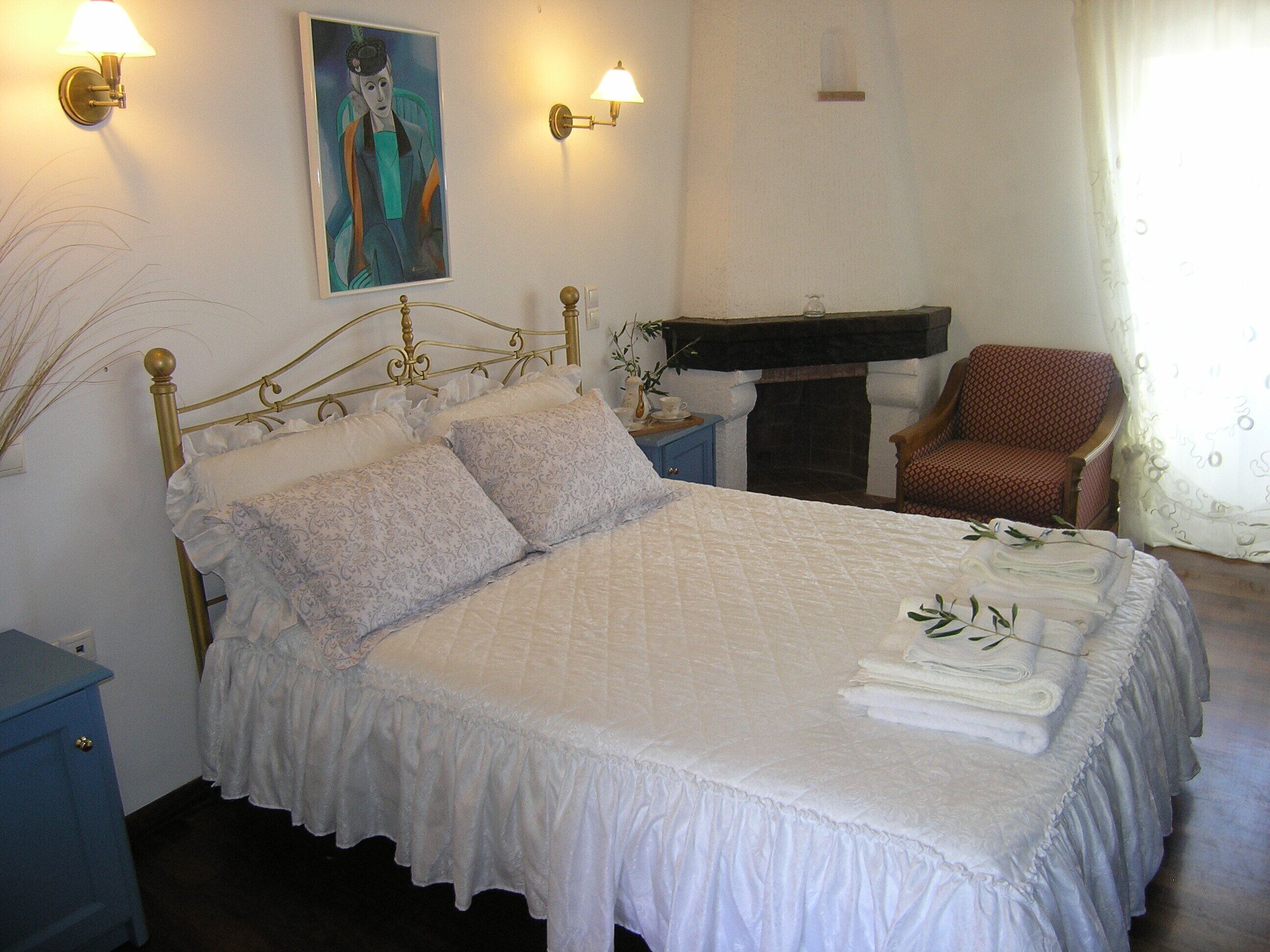 A neatly made bed with pillows, a blue nightstand, a painting on the wall, a chair, and a fireplace.