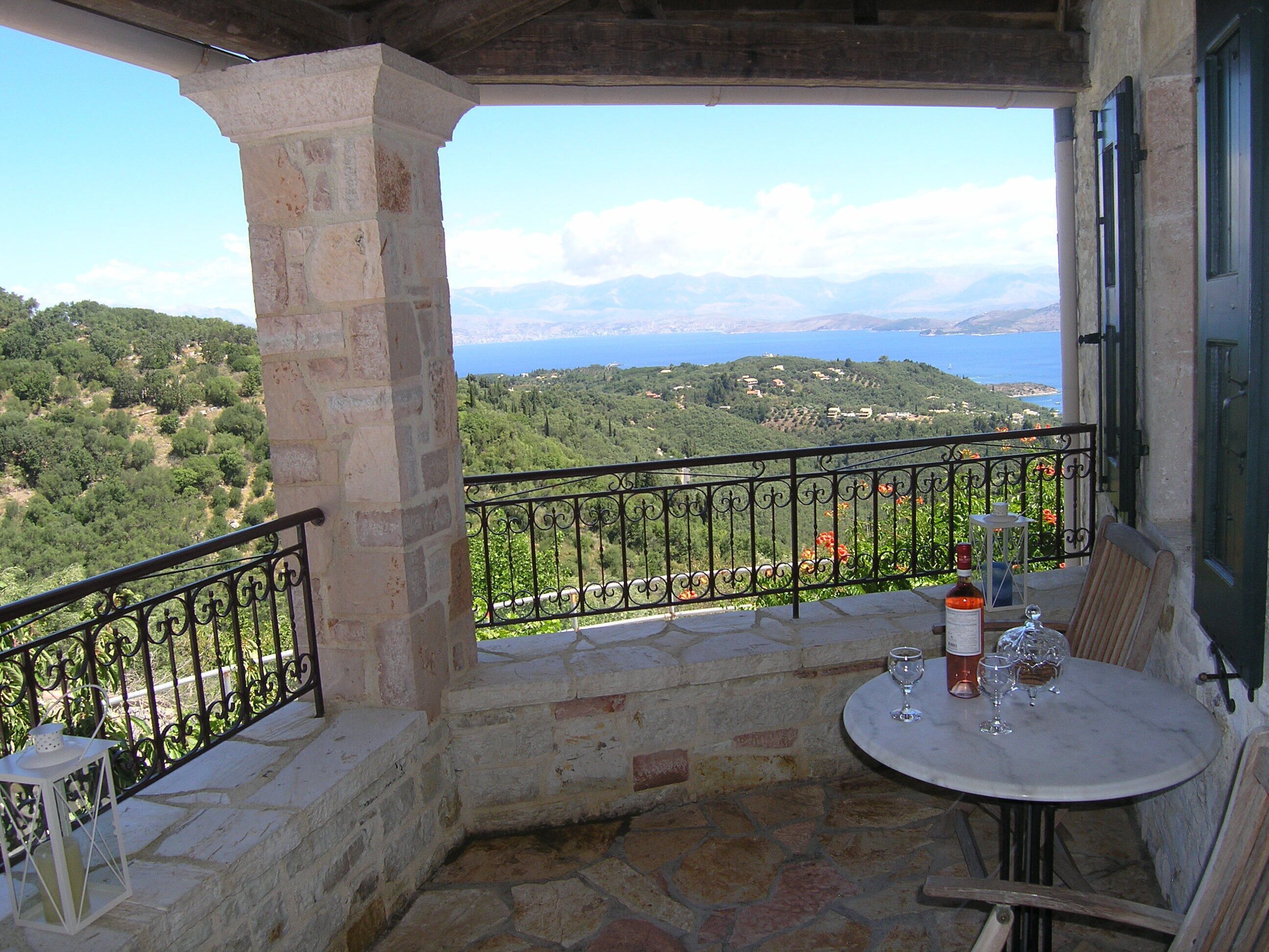 Un balcón con pared de piedra, barandilla metálica, una mesa con copas de vino y vista a un paisaje costero.