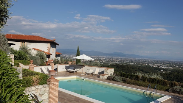 A swimming pool with lounge chairs and an umbrella, surrounded by stone walls and greenery, overlooking a cityscape. - Two-room apartment in old villa with pool located between Lucca and Florence (Pistoia)