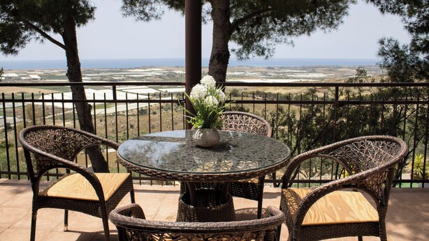 Una mesa redonda de vidrio con sillas de mimbre y un florero en un balcón con vistas a un paisaje pintoresco.