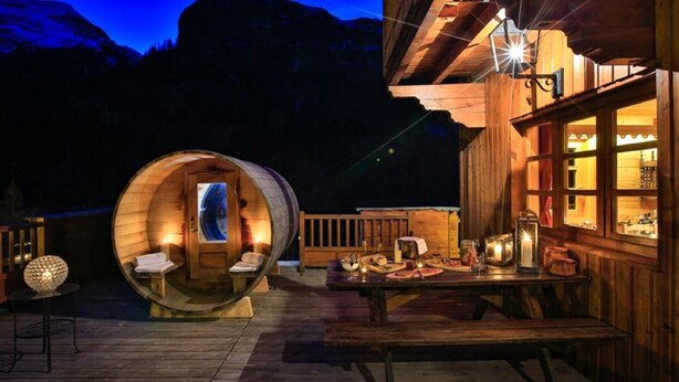 Una cabaña de madera con una habitación grande con forma de barril, una zona de comedor con una mesa para dos personas y vistas a montañas al fondo.