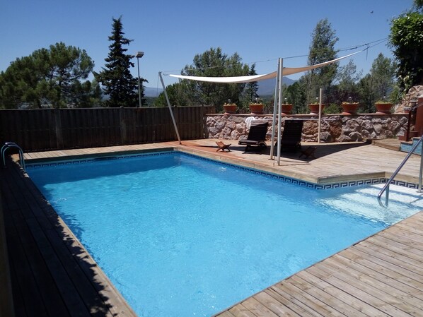 Una piscina rectangular con un deck de madera alrededor. Se observan sillones de descanso y una parrilla en el fondo.