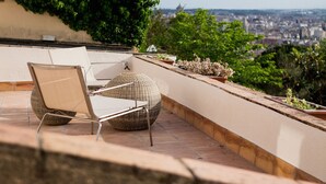 A rooftop terrace with a chair and a round wicker ottoman, potted plants, and a city view.