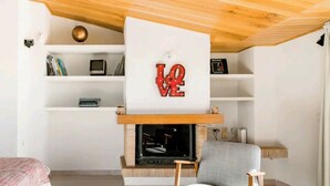 A cozy living room with a fireplace, a comfortable armchair, and a shelf with books and decorative items.
