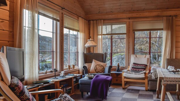 Un intérieur de chalet chaleureux avec des murs en bois, de grandes fenêtres, un coin salon confortable et un téléviseur.