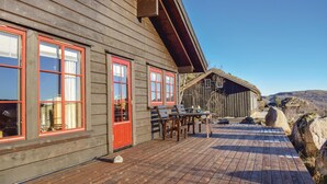 Une cabane en bois avec des fenêtres à cadre rouge, une terrasse dotée de mobilier d’extérieur et un paysage montagneux en arrière-plan.