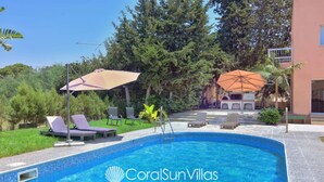 Una piscina con sillones de descanso y sombrillas, rodeada de vegetación y un edificio.