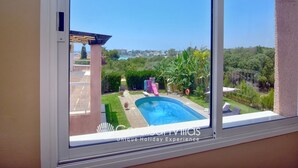 Vista desde una ventana de un área de piscina con cielo despejado, una pérgola y un jardín exuberante.