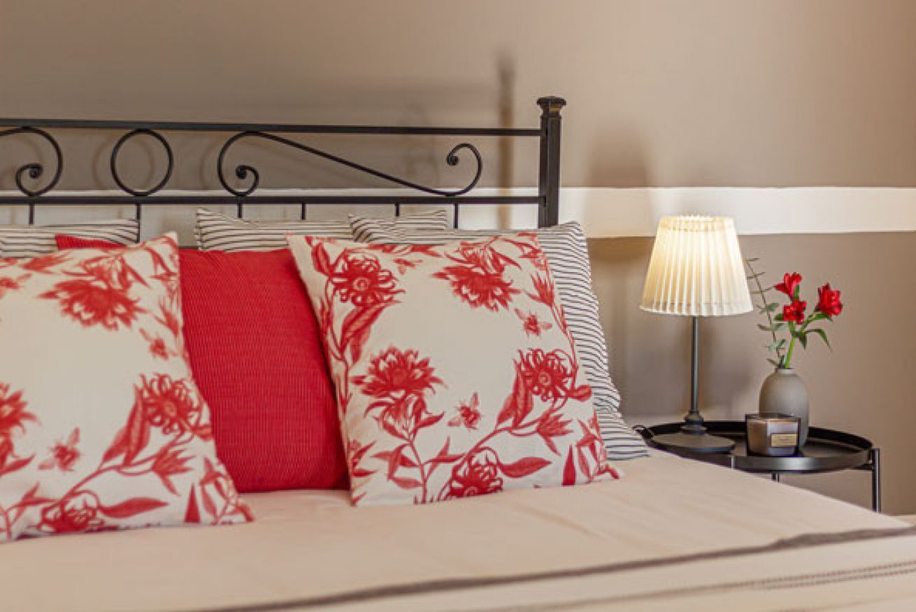 A neatly made bed with floral-patterned pillows and a striped pillow, a bedside table with a lamp and a vase of flowers, and a wall with a horizontal stripe.