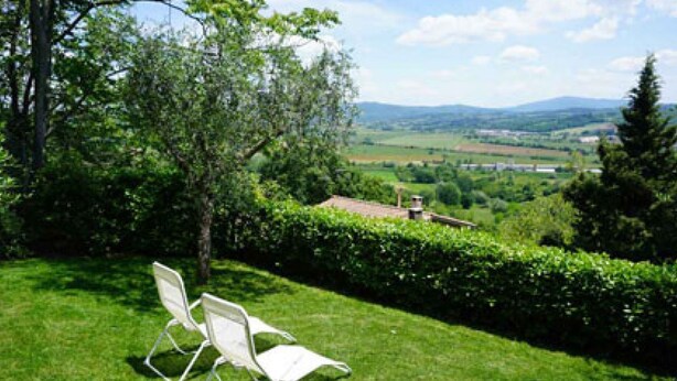Un espace extérieur verdoyant avec deux chaises blanches, une haie et une vue sur un paysage lointain comprenant des collines et un bâtiment.
