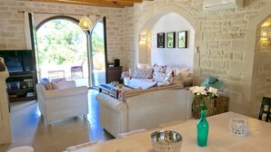 A cozy living room with a sofa, a dining table with a vase and candle, and a TV in the background. - Villa LaMori in a quiet location with a heatable pool and a beautiful garden (Nikiforos Fokas)