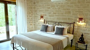 A bedroom with a stone wall, a bed with white bedding, patterned pillows, a small table with a statue, and a window with a curtain. - Villa LaMori in a quiet location with a heatable pool and a beautiful garden (Nikiforos Fokas)