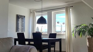 A spacious loft apartment with a high ceiling, a dining area with a table and chairs, a cozy seating area with a couch, and a large window providing a view of the city skyline. - Apartment Seeloft with balcony and direct lake view on the Möhnesee  (Möhnesee)