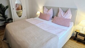 A neatly made bed with a patterned bedspread and pillows, a bedside lamp, a small wooden nightstand with a lamp, and a mirror on the wall. - Apartment Seeloft with balcony and direct lake view on the Möhnesee  (Möhnesee)