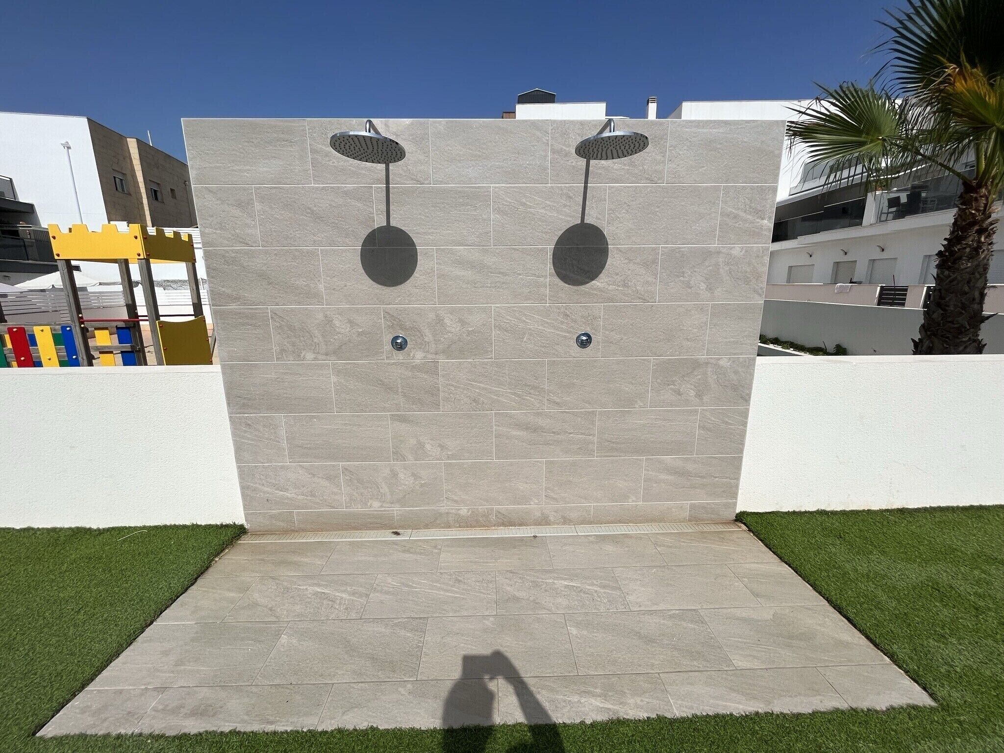 Un espace de douche extérieur moderne, doté de deux pommes de douche, d’un mur carrelé et d’une bordure d’herbe.
