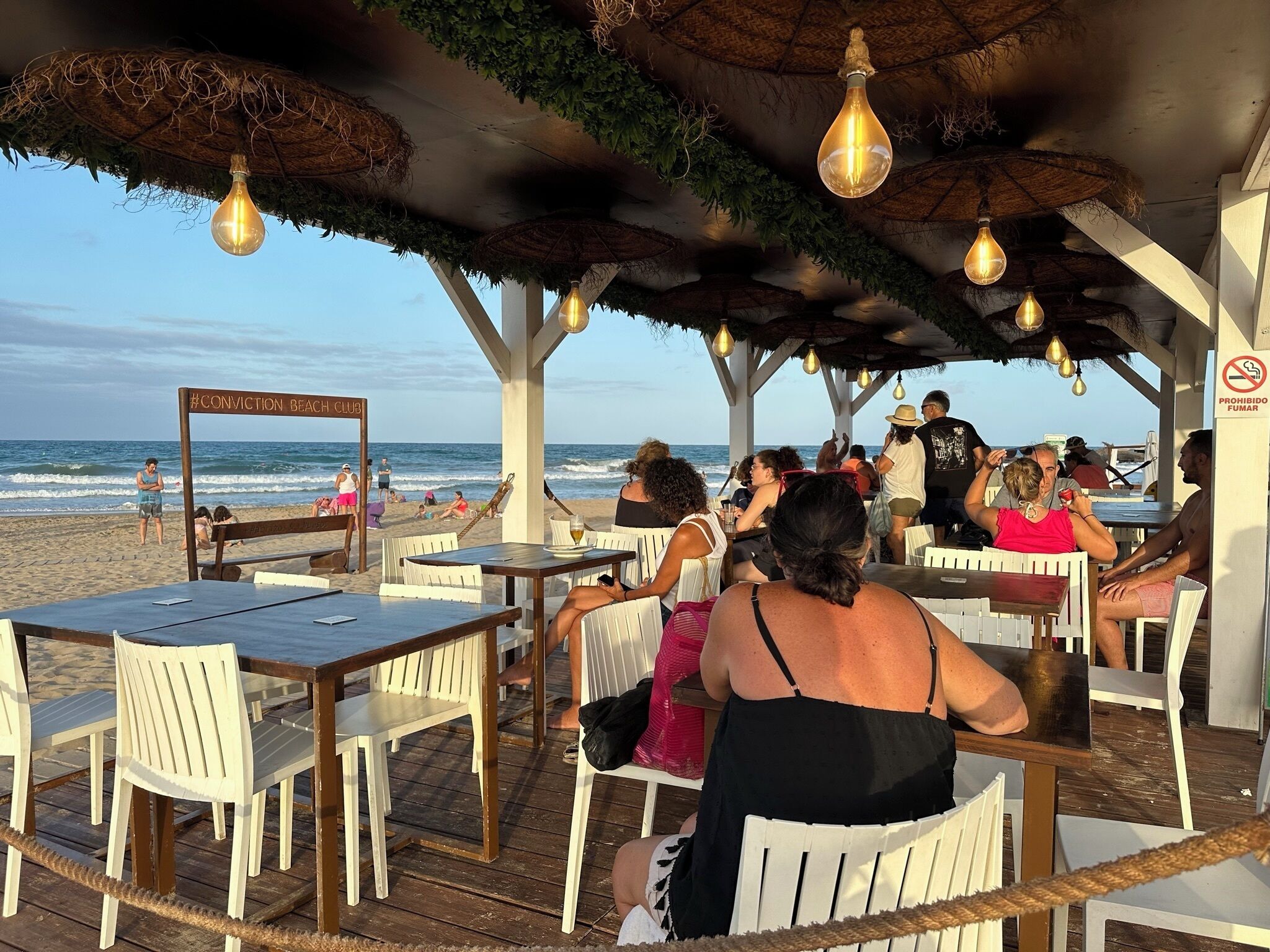 An outdoor seating area with tables and chairs, a beach view, and a sign reading 'Conviction Beach Club.'