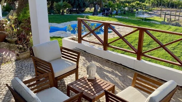 A patio with wooden furniture, a small table, and a view of a pool and greenery. - Da Silva Surfcamp Areia Branca (Lourinhã)