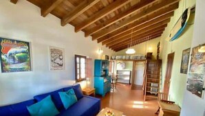 A cozy living room with a blue sofa, wooden beams, and a kitchen area in the background. - Da Silva Surfcamp Areia Branca (Lourinhã)
