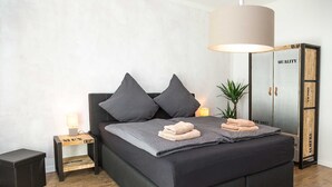 A modern bedroom with a large bed, dark bedding, two pillows, a bedside lamp, a small nightstand, and a wardrobe. - Apartment waterfront (Waren (Müritz))