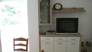 A room with a TV on a wooden cabinet, a clock, a small shelf with decorative items, and a window with a view of greenery. - Apartment in Lopar with Terrace, Air condition, WIFI (568-2) (Lopar)