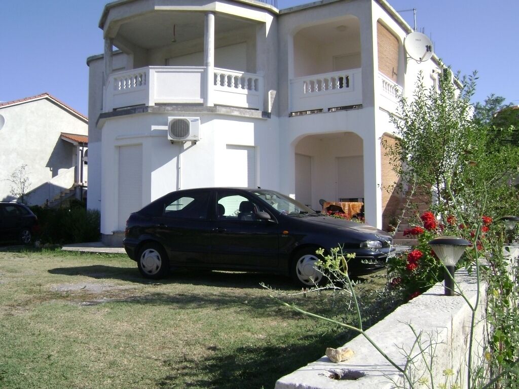 A two-story building with a balcony, a parked car, and a garden with red flowers.
