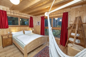 A wooden cabin bedroom with a bed, nightstands, a hanging chair, and a wooden ladder.