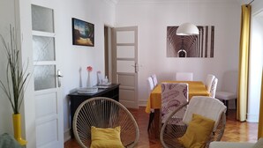 Une pièce avec des chaises en osier, une table à manger recouverte d’une nappe jaune, un canapé blanc, un tableau et un luminaire suspendu.
