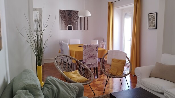 Un salon avec un canapé blanc, une table à manger avec des chaises, un vase avec des plantes et des œuvres d’art accrochées aux murs.
