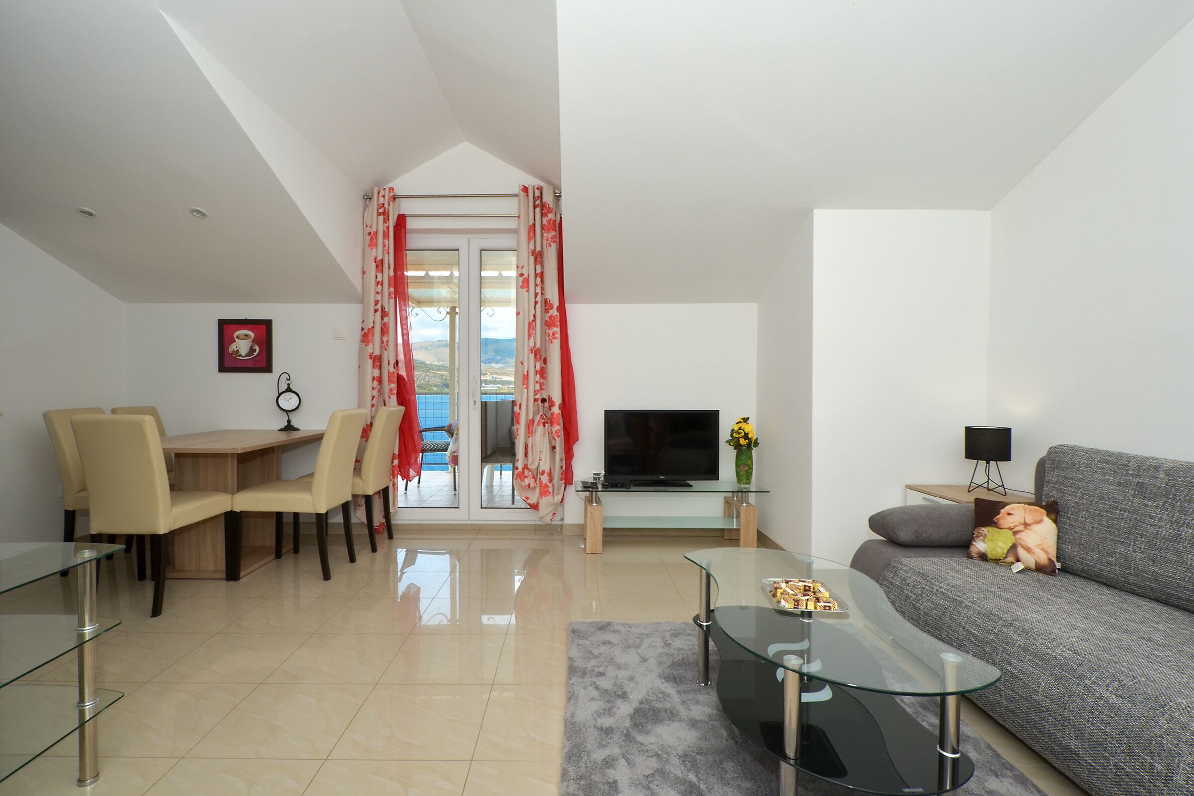 A modern living room with a glass dining table, a sofa, a TV, and a balcony with red curtains.