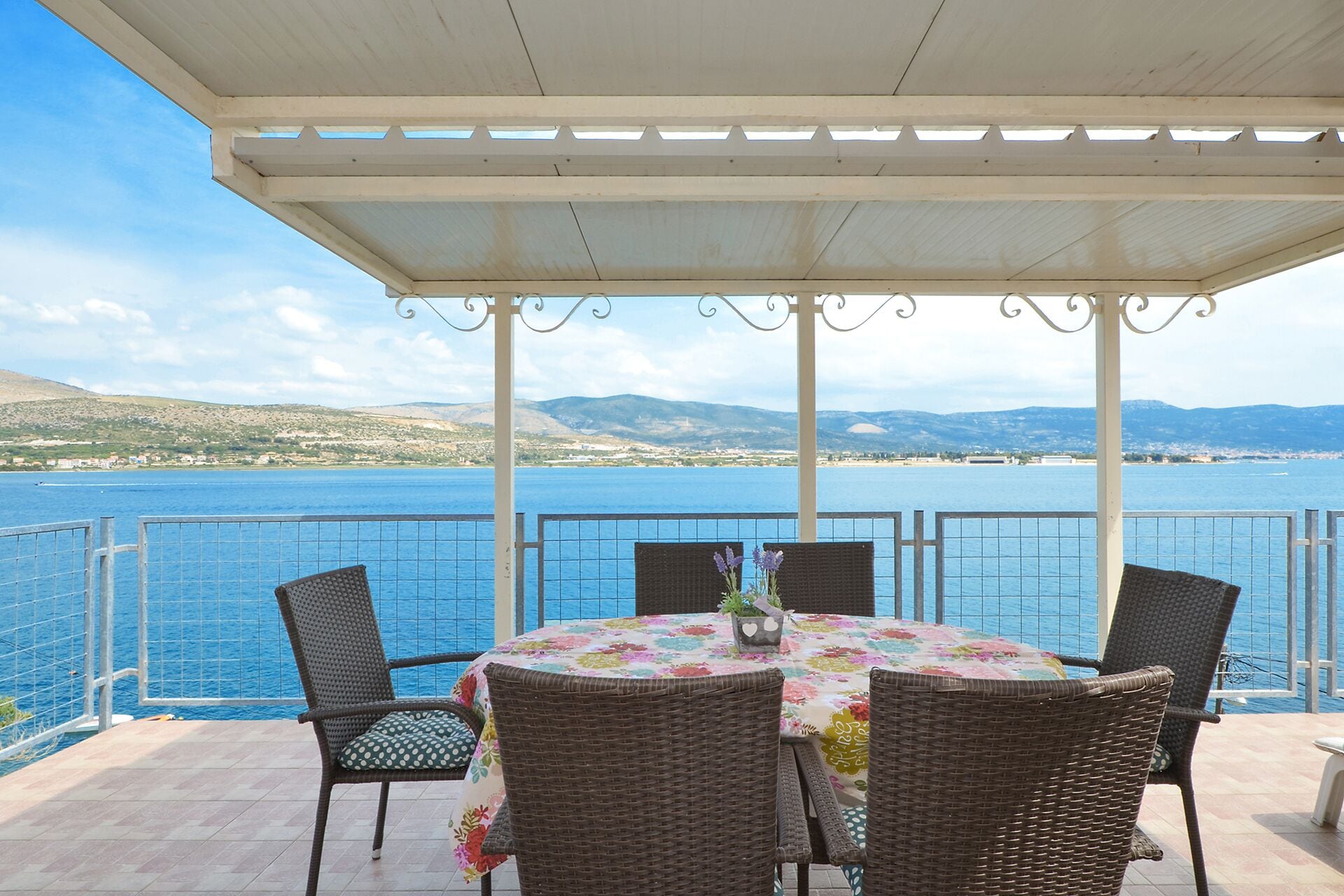 A covered outdoor seating area with a table and chairs, overlooking a body of water and distant hills.