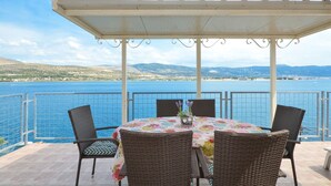 Area salotto esterna coperta con tavolo e sedie, affacciata su uno specchio d'acqua e dolci colline.